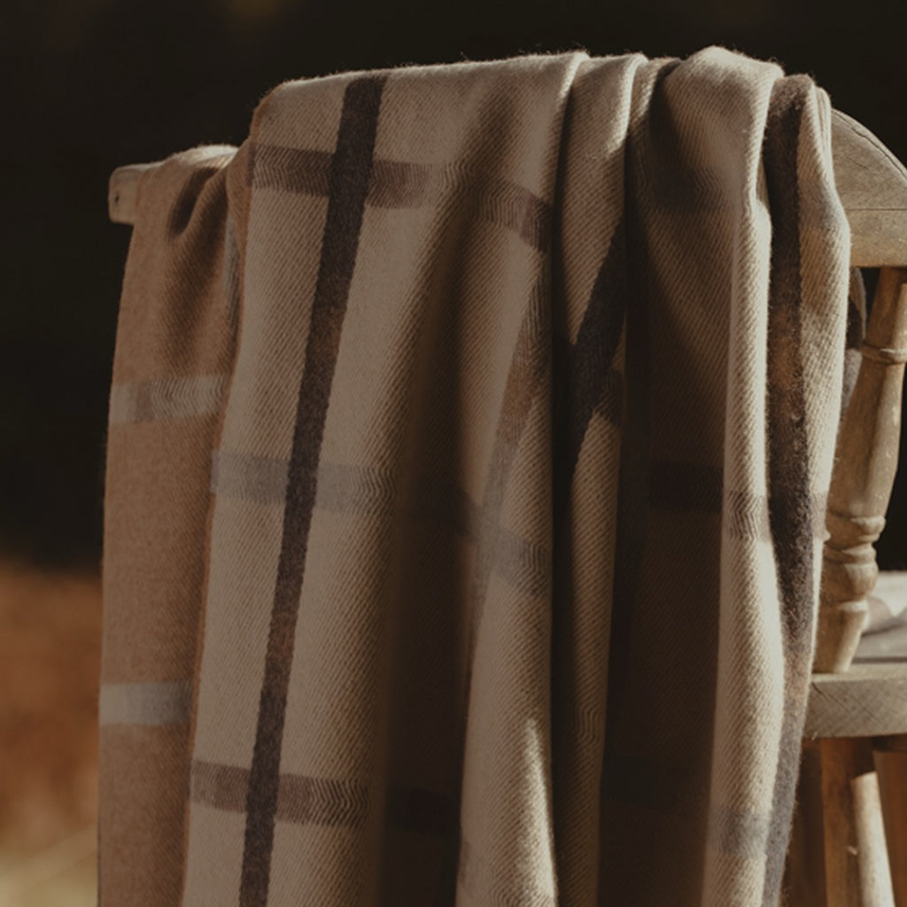A beige and brown plaid merino throw blanket draped over a rustic wooden chair in warm, dark lighting.