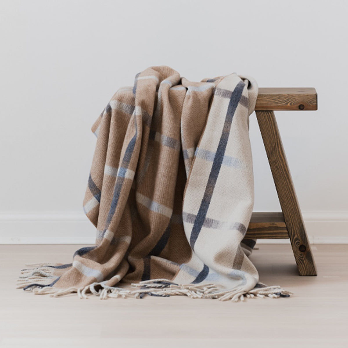 A soft woven reversible throw blanket with a beige, cream, and brown plaid pattern and tasseled fringe, draped over a rustic wooden stool against a neutral light grey wall.