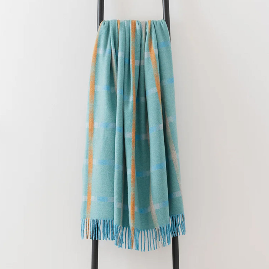 A soft woven throw blanket in a light aqua blue colour, featuring vertical orange stripes  and pale blue and cream horizontal stripes and a subtle plaid pattern, draped over a black metal stand against a plain white wall.