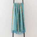 A soft woven throw blanket in a light aqua blue colour, featuring vertical orange stripes  and pale blue and cream horizontal stripes and a subtle plaid pattern, draped over a black metal stand against a plain white wall.