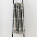 A gray plaid throw blanket with cream and white large overchecks and fringed edges, draped over a black metal ladder against a plain white wall.