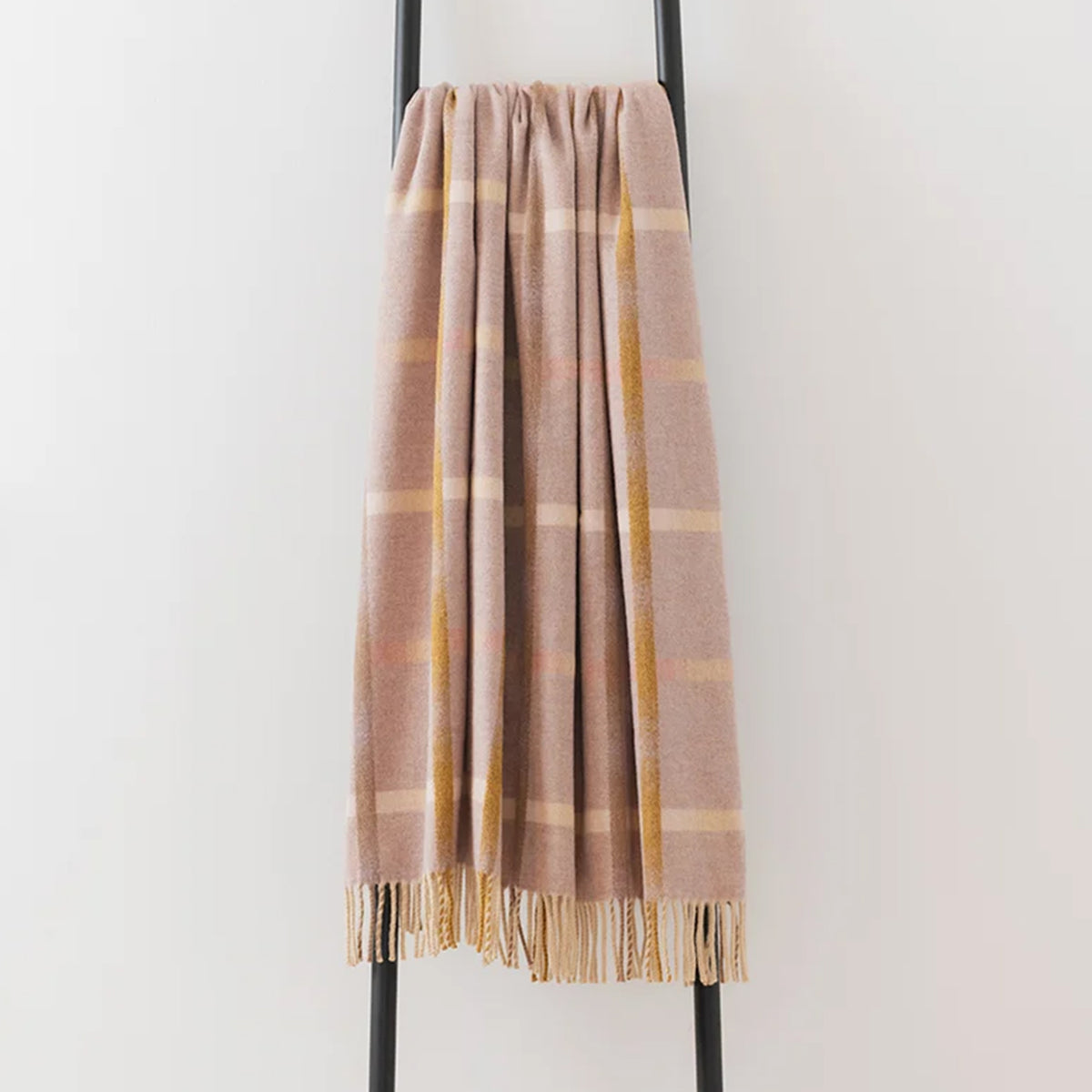 A soft woven throw blanket draped over a black metal stand, featuring a muted pink/beige base with warm orange and cream stripes and fringed edges.