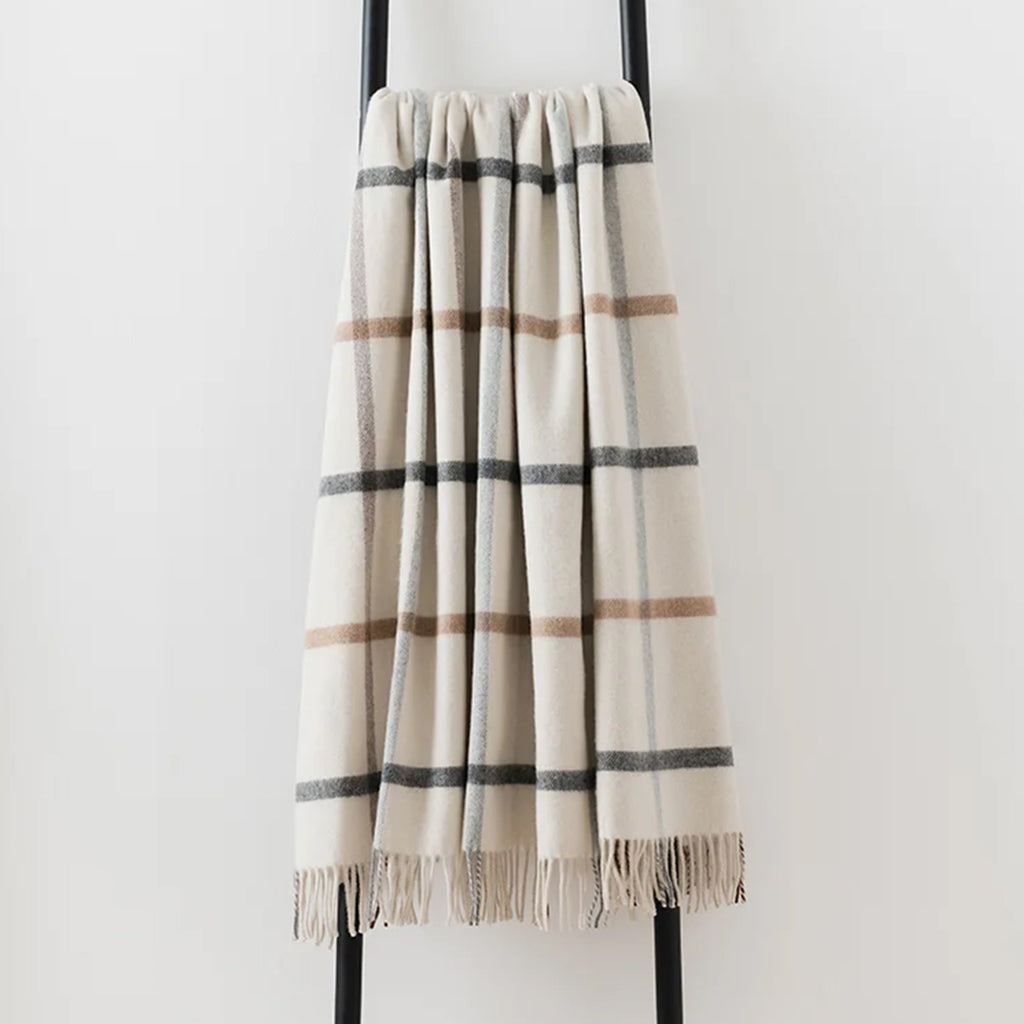 A cream-colored plaid merino  throw blanket with fringe, draped over a black metal stand against a plain white wall.