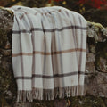 A cream-colored woven merino throw blanket with a brown and grey plaid pattern and fringed edges, draped over a rustic stone wall outdoors.