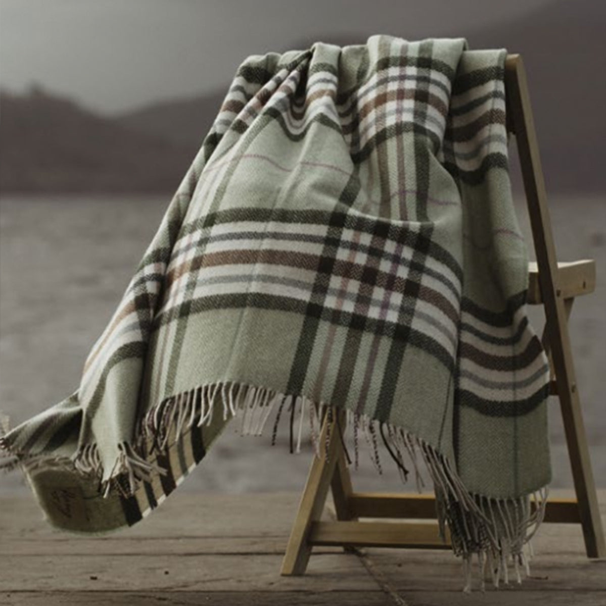 A large jade green herringbone wool blanket with brown and purple stripes, hanging outdoors on a chair next to the loch