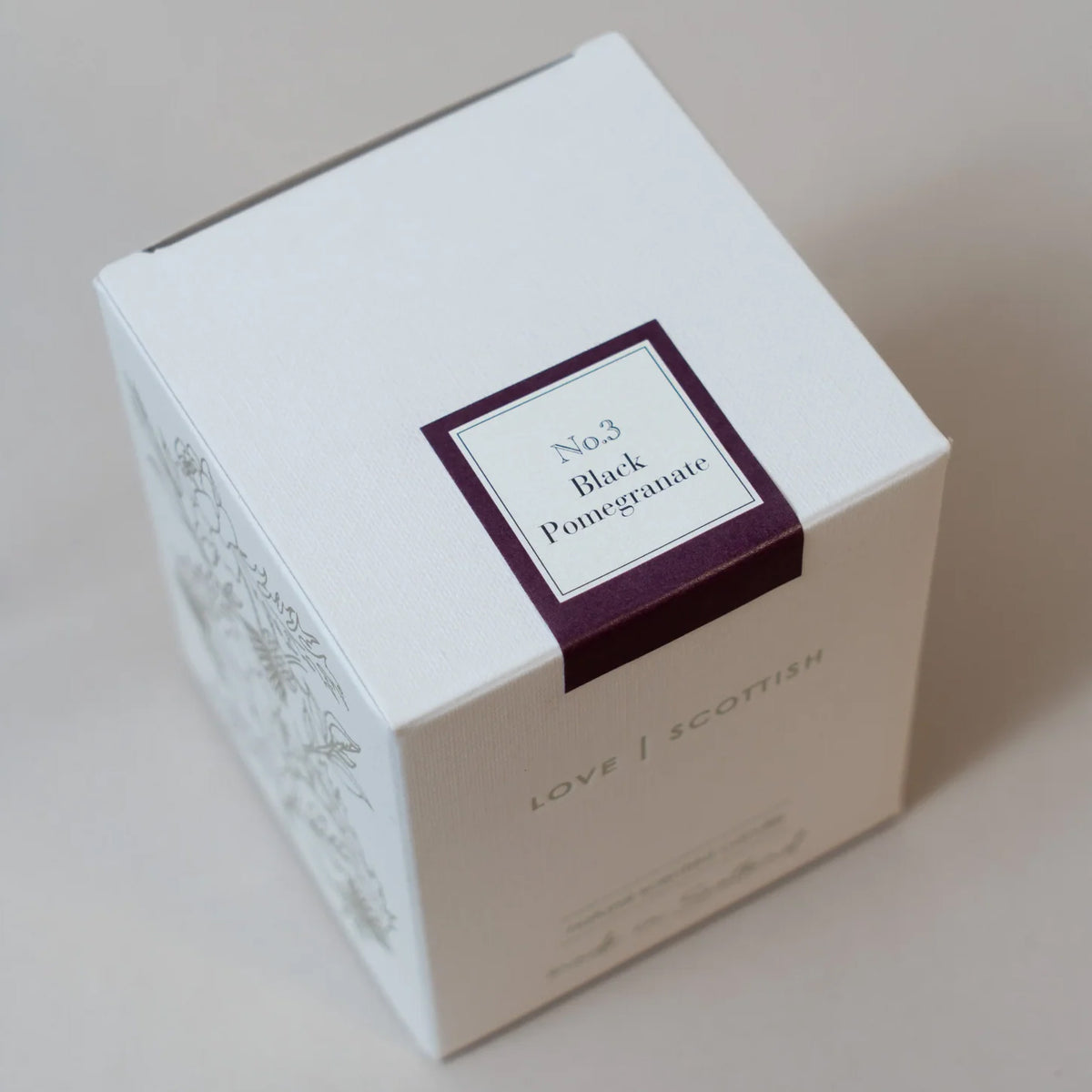 A white square product box with a dark purple label reading 'No.3 Black Pomegranate Candle' and 'LOVE SCOTTISH' printed on the side, photographed on a plain light beige background.