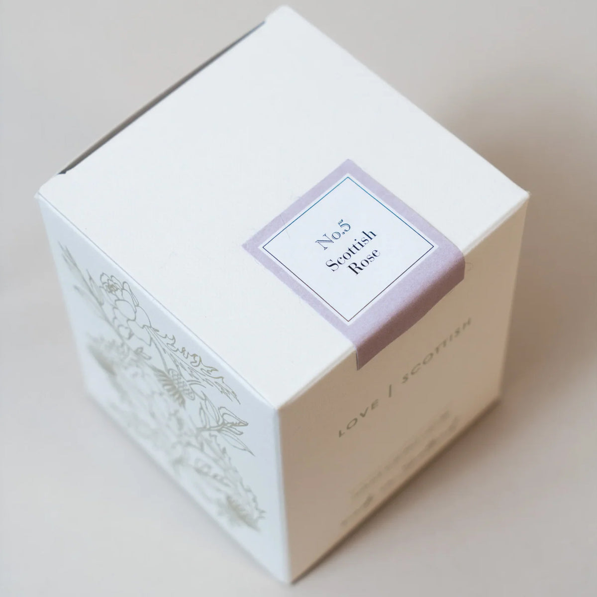 A square white product box with a lavender label reading 'No.5 Scottish Rose Candle' and delicate floral line art on the side, photographed on a warm beige background.