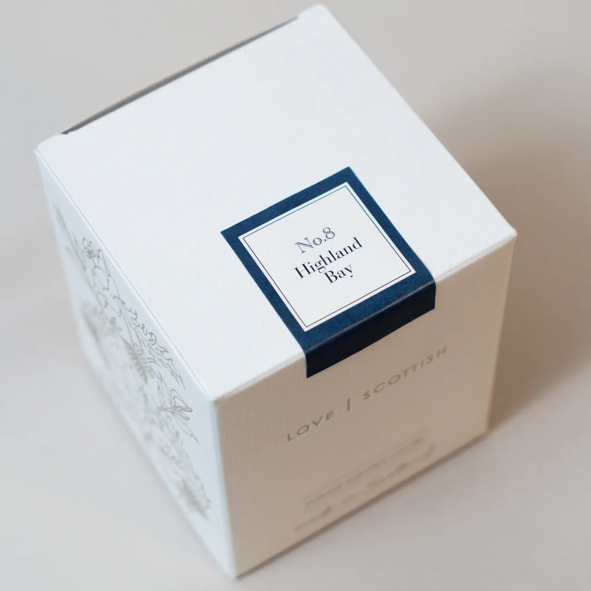 A white square product box with a navy blue label reading 'No.8 Highland Bay Candle' and 'LOVE SCOTTISH' printed on the side, photographed on a light neutral background.
