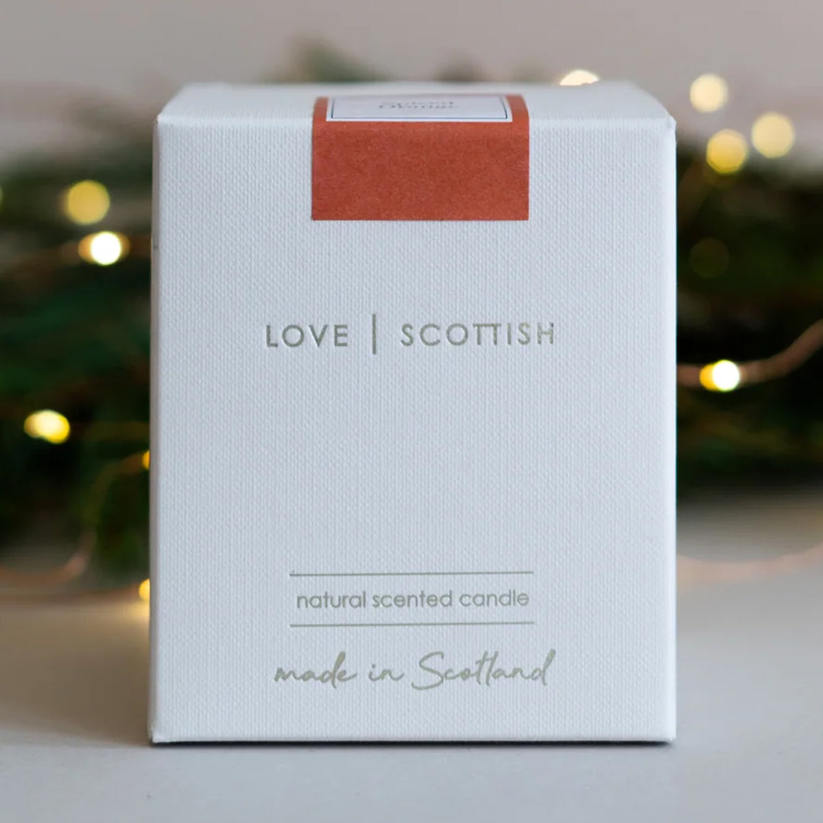 A white square product box for a 'Spiced Orange' scented candle by 'Love Scottish'.  Featuring a magenta label on the top face and delicate line art on the side, set against a softly blurred background with warm fairy lights.