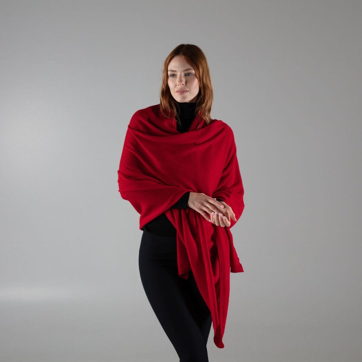 A woman modeling a vibrant red shawl or wrap draped over her shoulders, paired with a black turtleneck and black leggings, set against a solid gray studio background.