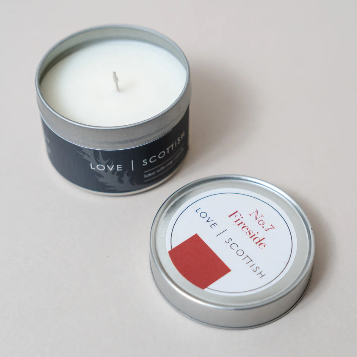 An open silver tin candle with white wax sits next to its matching lid on a light beige background. The label reads 'LOVE SCOTTISH' , Fireside Scent