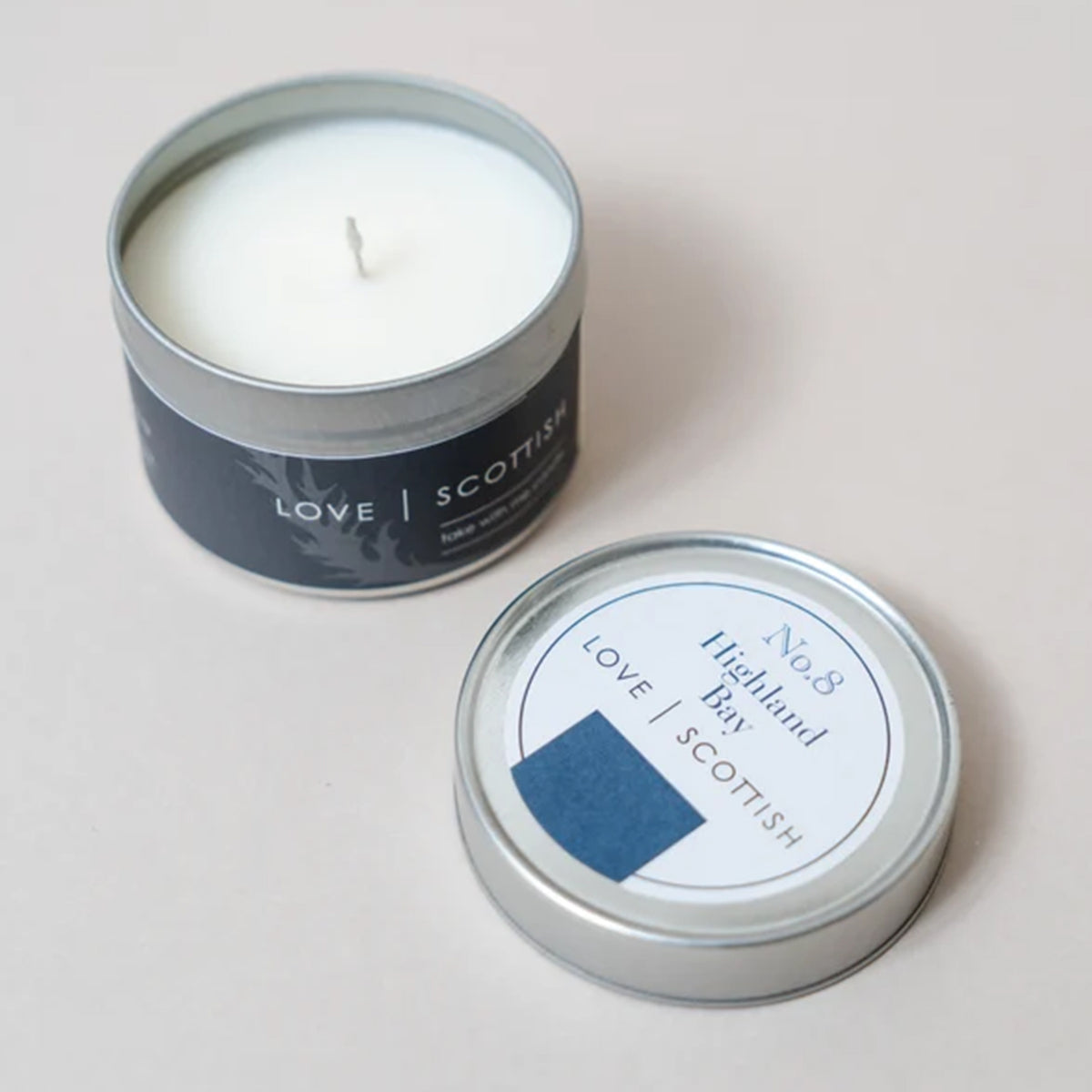 A white wax candle in a round silver metal tin with a dark navy label reading 'LOVE SCOTTISH', scent Highland Bay. The tin is shown open with the candle inside, and the matching lid placed in front on a light beige background.