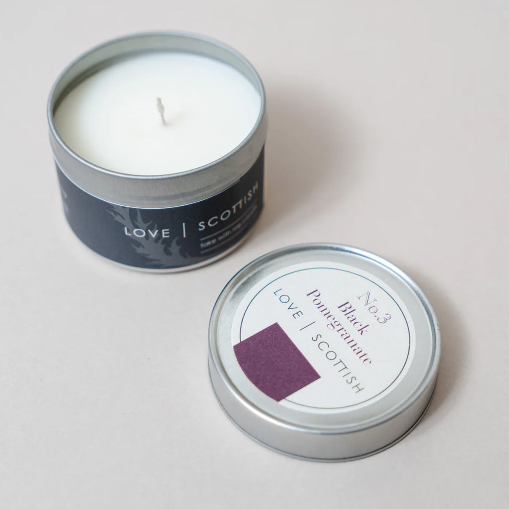 An open silver tin travel candle with white wax sits next to its matching lid on a light beige background. The label reads 'LOVE SCOTTISH' , black pomegranate scent
