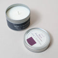 An open silver tin travel candle with white wax sits next to its matching lid on a light beige background. The label reads 'LOVE SCOTTISH' , black pomegranate scent