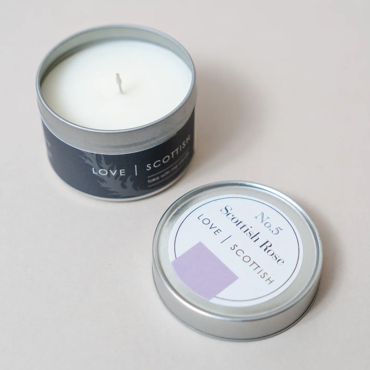 An open silver tin candle with white wax sits next to its matching lid on a light beige background. The label reads 'LOVE SCOTTISH' , Scottish Rose Scent