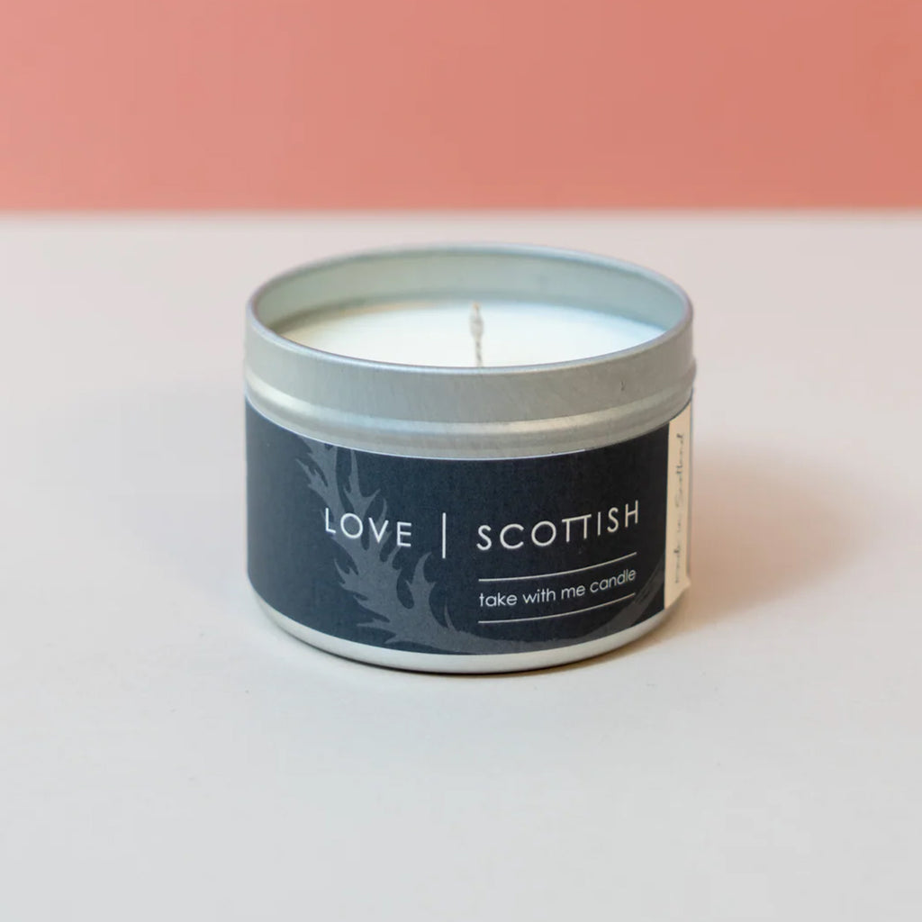 A single-wick candle in a round silver metal tin with a dark navy label reading 'LOVE SCOTTISH' and 'take with me candle.' The candle is photographed on a white surface against a soft pink background.