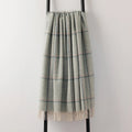 A soft woven plaid throw blanket in green hue with cream and blue stripes, draped over a black metal stand against a plain light gray wall.