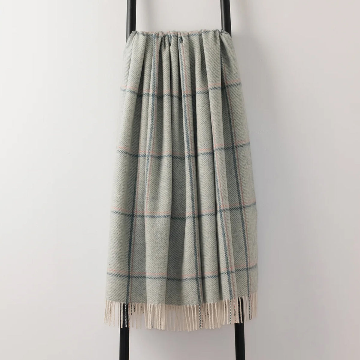 A soft woven plaid throw blanket in green hue with cream and blue stripes, draped over a black metal stand against a plain light gray wall.