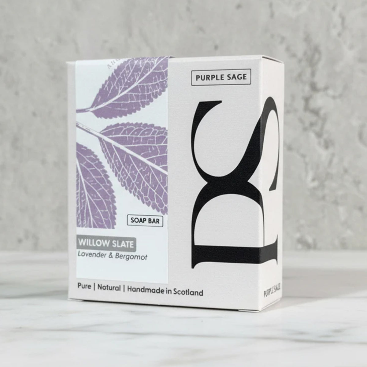 A boxed soap bar package standing upright on a light marble surface against a textured light gray wall. The box is white with large black lettering and a purple leaf illustration.