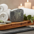 A rectangular bar of soap with a dark gray and pale green marbled swirl pattern, displayed on a wooden tray with white towels, smooth stones, and eucalyptus leaves. In the background, blurred white candles glow with a warm, spa-like ambiance.