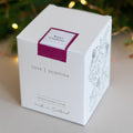 A white square product box for a 'Winter Cranberry' scented candle by 'Love Scottish'.  Featuring a magenta label on the top face and delicate line art on the side, set against a softly blurred background with warm fairy lights.