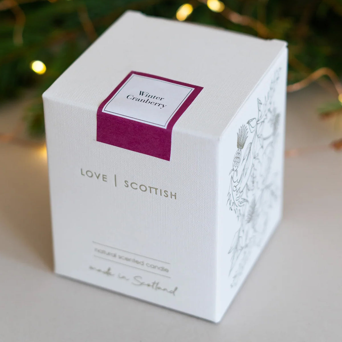 A white square product box for a 'Winter Cranberry' scented candle by 'Love Scottish'.  Featuring a magenta label on the top face and delicate line art on the side, set against a softly blurred background with warm fairy lights.