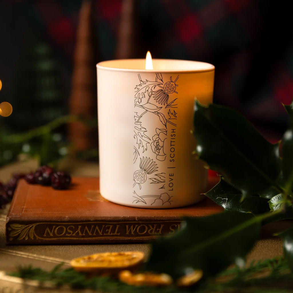 A lit white candle with a vertical line-art illustration of a thistle and the words 'LOVE SCOTTISH' on the front, styled on a vintage book with holly leaves and dark berries in a warm, festive setting.