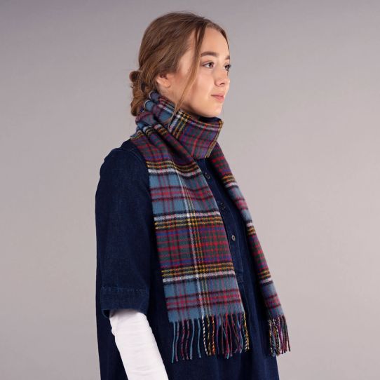 A woman in profile wearing a navy blue denim dress and a thick plaid scarf in shades of blue, red and yellow, set against a plain grey studio background.