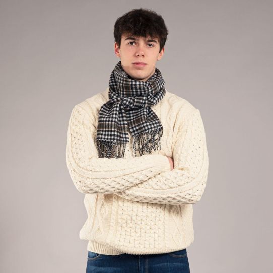 A close up of a man with crossed arms wearing a knitted cream jumper with a tartan scarf looped around his neck.