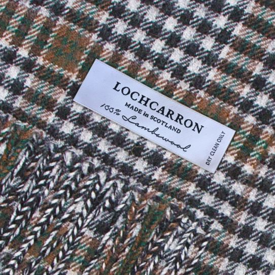 Close-up of a textured woven fabric with a white label reading 'LOHCARRON MADE IN SCOTLAND 100% Lambwool DRY CLEAN ONLY'. The fabric has a neutral tartan design in brown, green, black, and off-white.