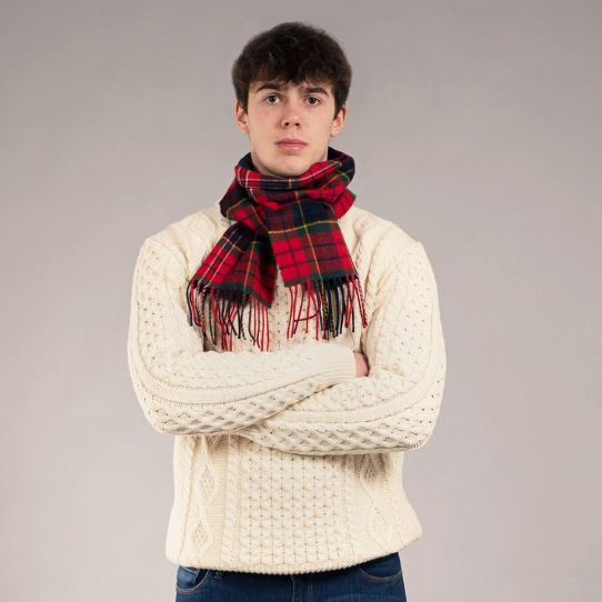 A young man wearing a cream cable-knit sweater and a red tartan plaid scarf with black and green stripes, standing against a neutral gray studio background.