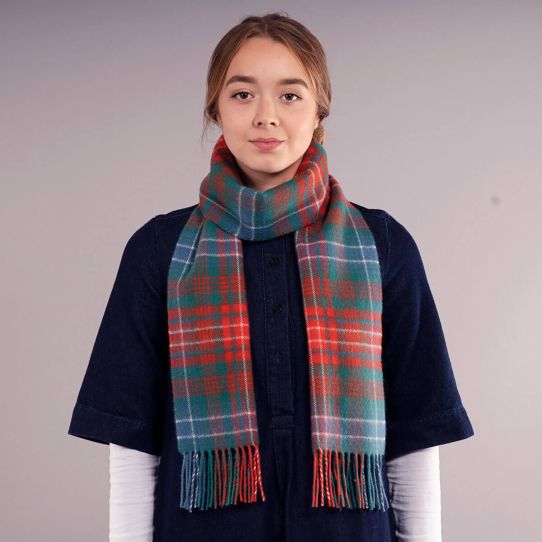A young woman wearing a navy denim blue top and a red, green, and blue plaid scarf with fringe, standing against a plain light grey background.
