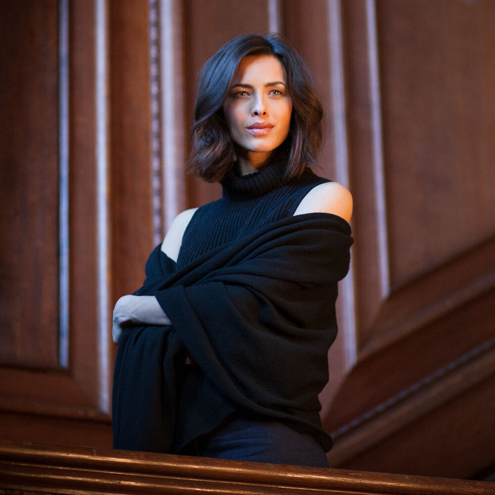 A woman with shoulder-length dark brown hair stands in front of a dark wood-paneled wall, wearing a black off-the-shoulder knit top with a draped black shawl or wrap.