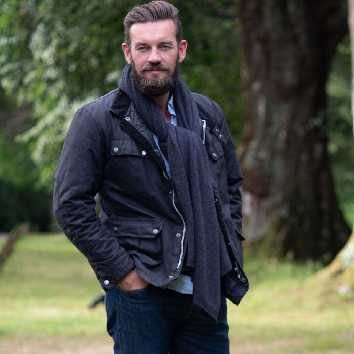 A man wearing a black utility jacket, dark grey knit cashmere scarf, and blue jeans, standing outdoors in a park with trees in the background.