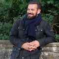 A smiling man with a beard wearing a black waxed jacket and a navy blue cashmere scarf, leaning against a stone wall with green foliage in the background.