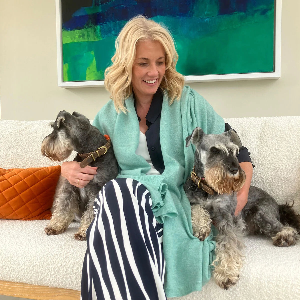 A woman sitting on a white sofa with two gray dogs, wrapped in a light teal jade wrap. She is wearing a black and white striped skirt and a black top.