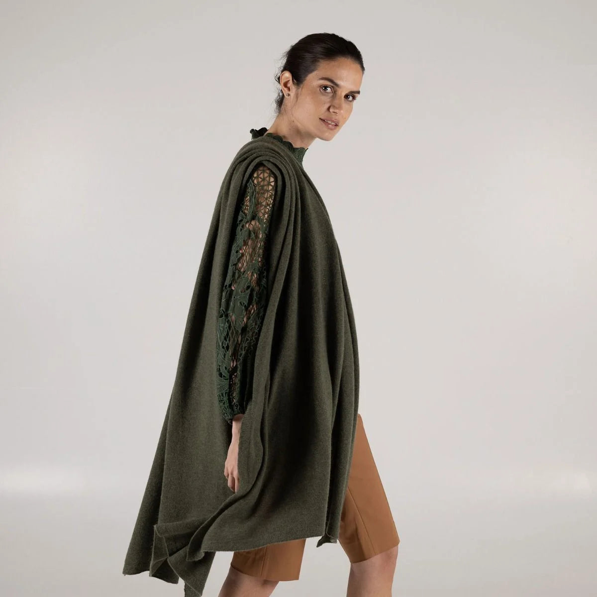 A woman modeling a dark olive green draped wrap, paired with brown shorts, against a plain light gray studio background.