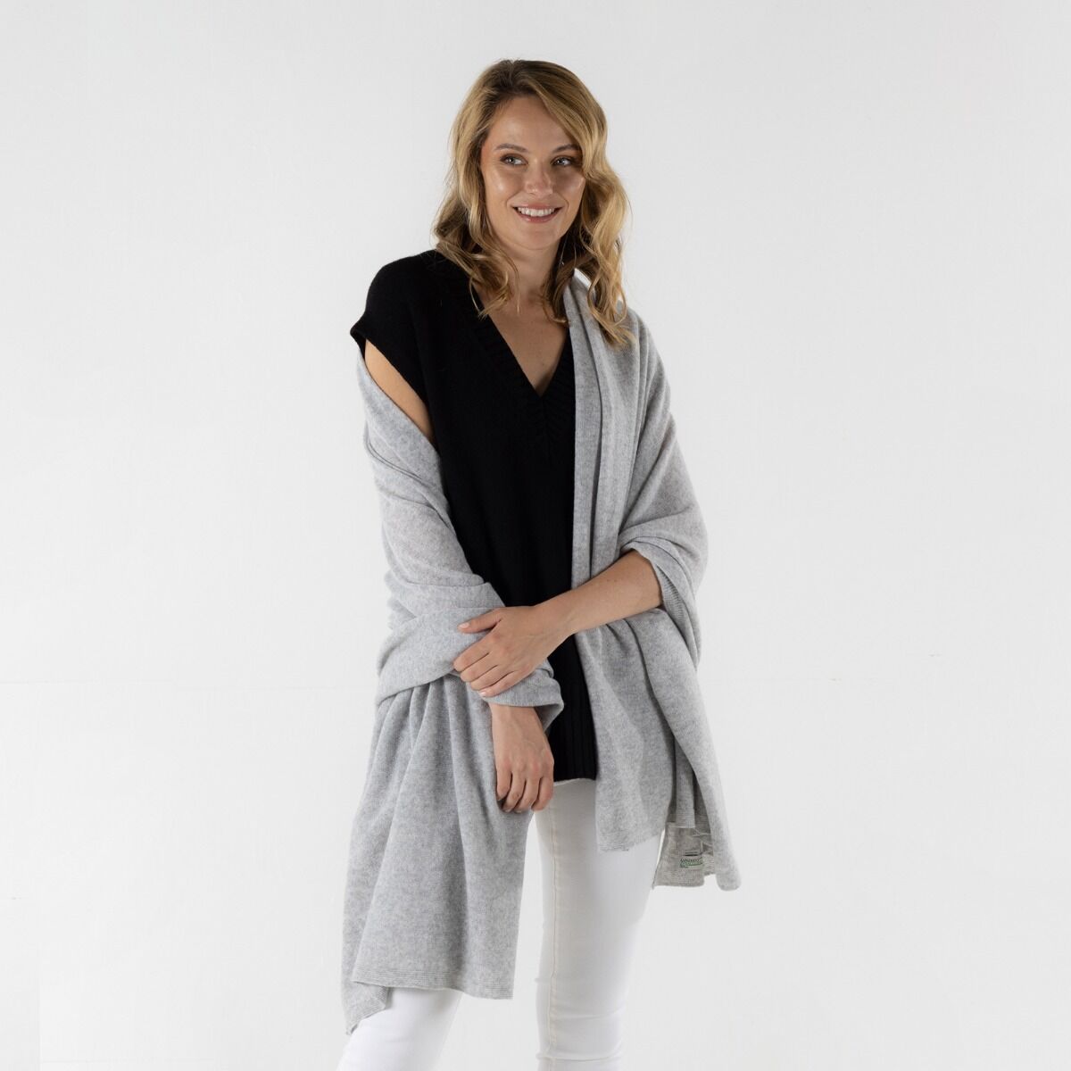 A woman modeling a light gray knit wrap or shawl draped over a black top and white pants, photographed against a clean white studio background.