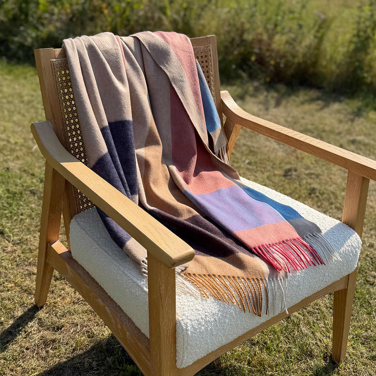 A multicolor beige patchwork wool throw blanket with fringe is draped over a wooden armchair with a cream bouclé seat cushion, set outdoors on a grassy lawn.
