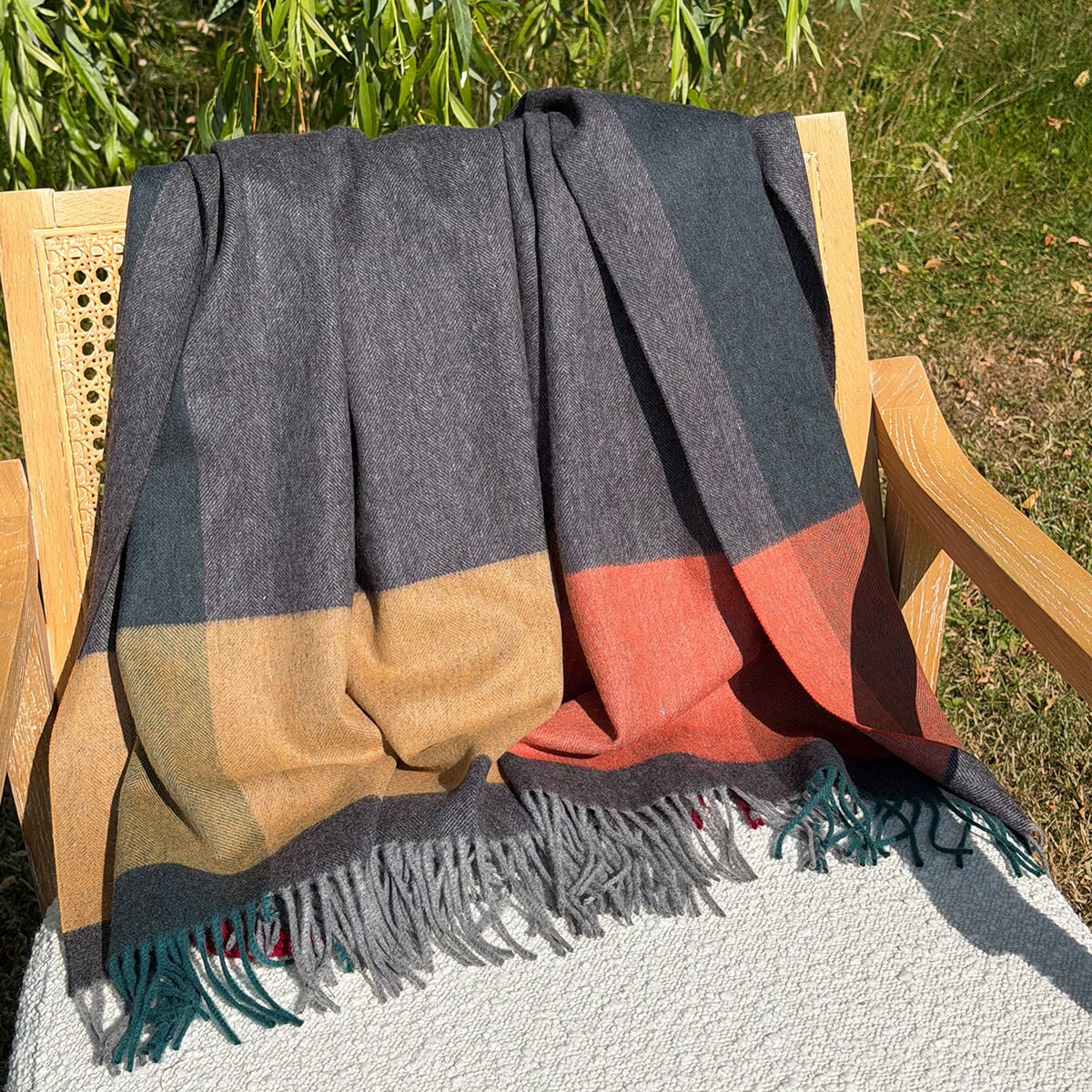 A large grey woven wool throw blanket with a block pattern in charcoal gray, mustard yellow, rust orange, and teal fringe, draped over a wooden chair in an outdoor garden setting.