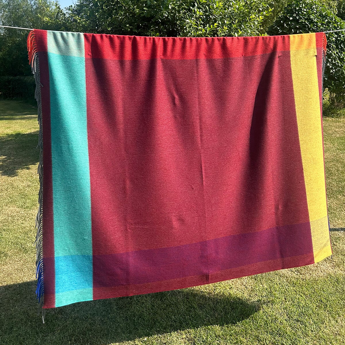 A large red woven wool blanket or throw with bold color-blocked stripes, hanging outdoors on a clothesline over a grassy lawn with trees in the background.