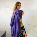 A woman with long light brown hair stands in front of a white marble wall, wearing a purple knit wrap draped over her shoulders. She is also wearing a black dress with a colourful floral pattern.