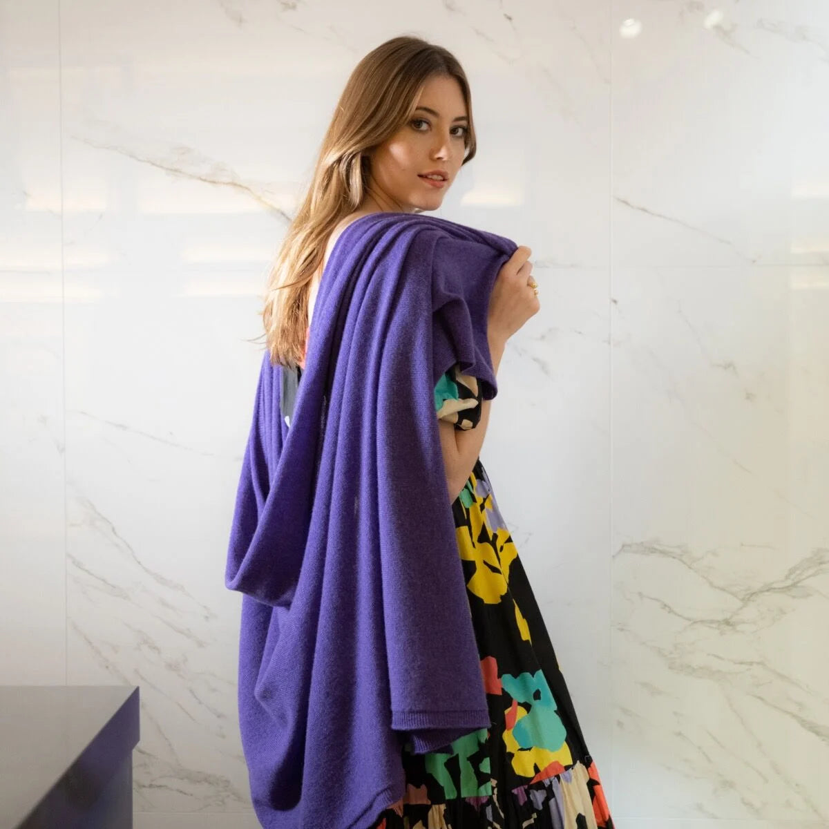 A woman with long light brown hair stands in front of a white marble wall, wearing a purple knit wrap draped over her shoulders. She is also wearing a black dress with a colourful floral pattern.