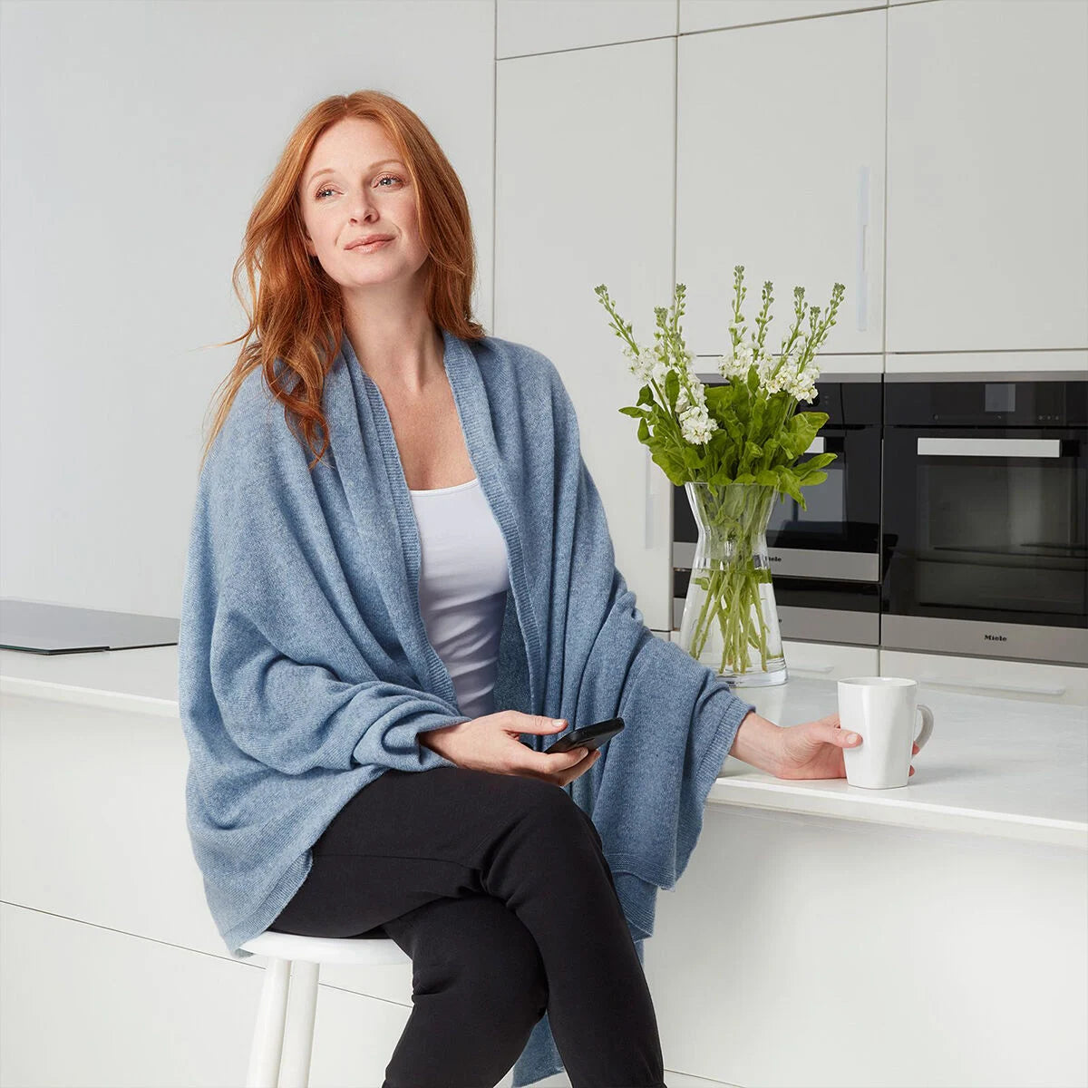 A woman with red hair sits on a white stool in a modern kitchen, wearing a light blue knit shawl over a white top and black pants. She holds a smartphone and reaches toward a white mug on the counter, with a vase of flowers and kitchen cabinets in the background.