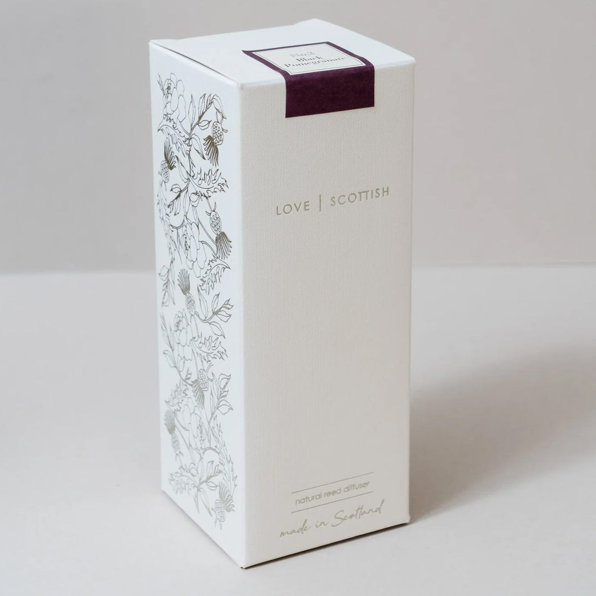 A tall white rectangular product box with botanical line drawings on the side, the words 'LOVE SCOTTISH' on the front, and a dark purple band near the top, inside a reed diffuser, pomegranate scent. photographed on a light neutral background.