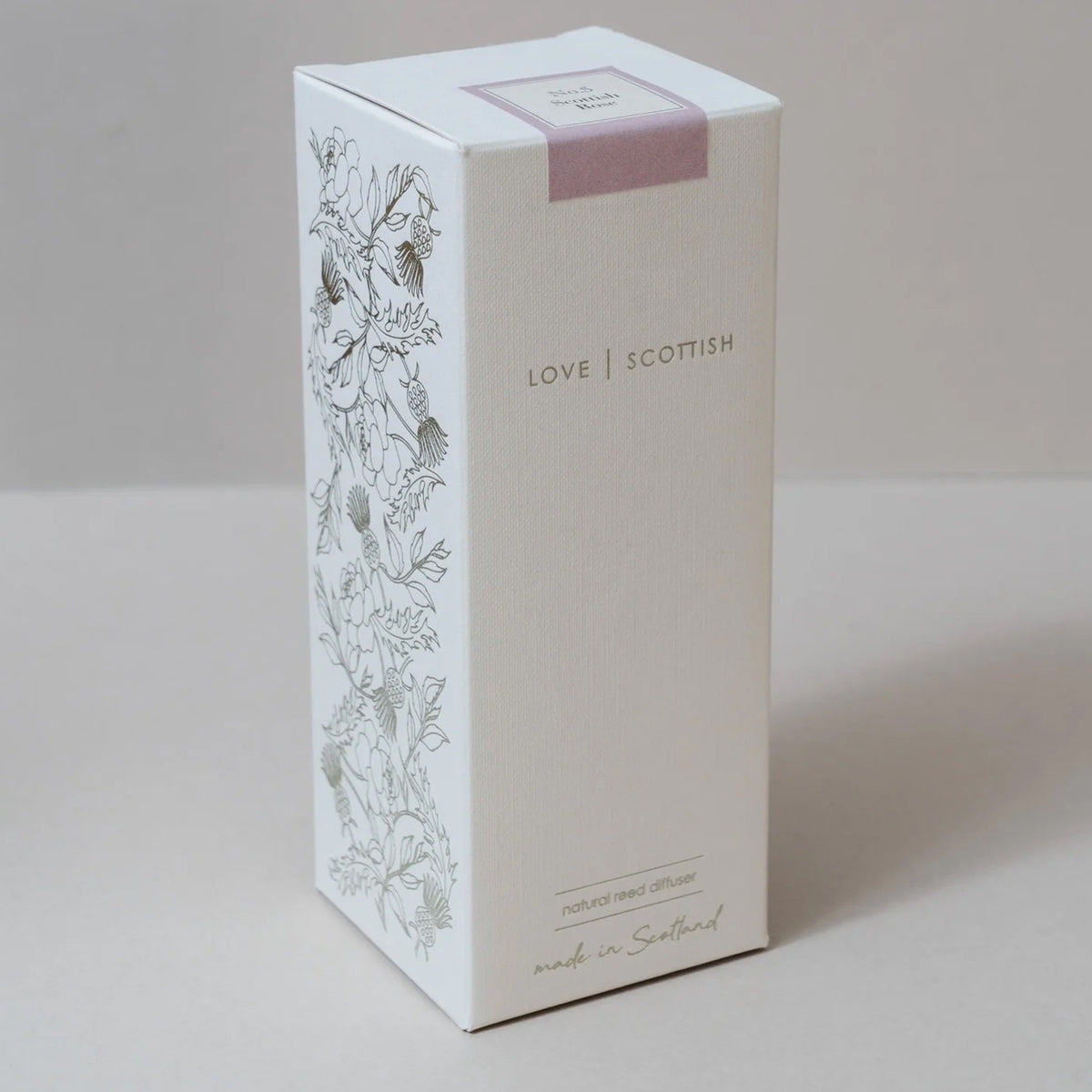 A tall white rectangular product box with floral line art on the side panel, a pale pink label on top, and the text 'LOVE SCOTTISH' printed on the front, reed diffuser in a Scottish rose scent. photographed on a light neutral background.
