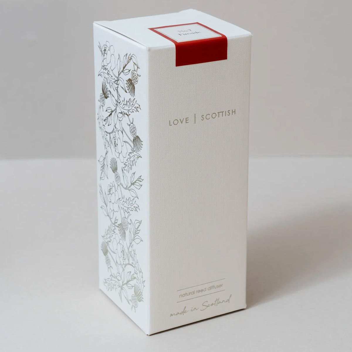A tall white rectangular product box with floral line art on the side, the words 'LOVE SCOTTISH' on the front, and a small red band around the top, inside a reed diffuser with a fireside scent.  photographed on a light neutral background.