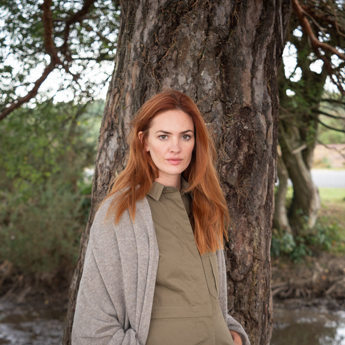 A woman with long red hair stands outdoors leaning against a large tree trunk, wearing an olive green top and a brown wrap.