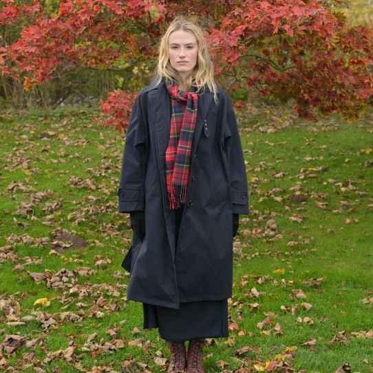 A woman stands outdoors in a grassy area with fallen leaves and red autumn foliage in the background, wearing a long dark navy coat over a dark dress, a red plaid scarf, and brown lace-up boots.