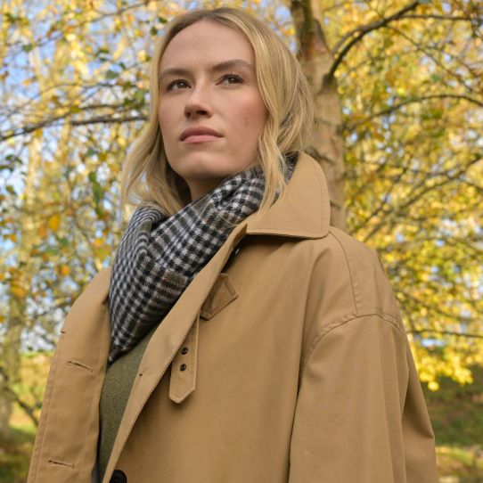 A close up of a model in the forest in a trench coat with a classic tartan scarf around her neck in neutral colours.